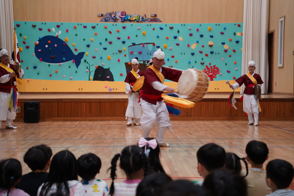 합굿마을 풍물마당놀이 해피 공연단이 북춤을 추는 모습 배경에 보랏빛 고개가 그려진 클라이밍 벽이 있는 모습