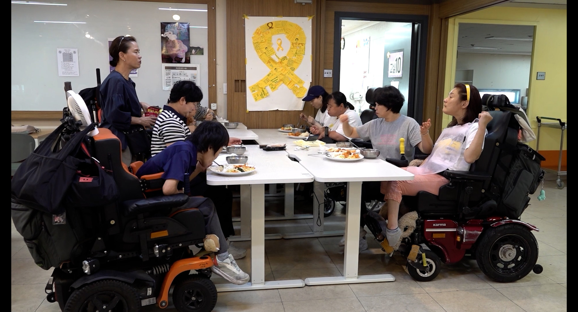 마스크를 쓴 두 사람이 앉아있다. 오른쪽 사람은 녹음용 마이크 앞에 앉아 헤드셋을 끼고 있고, 왼쪽 사람은 어딘가를 가리키고 있다.