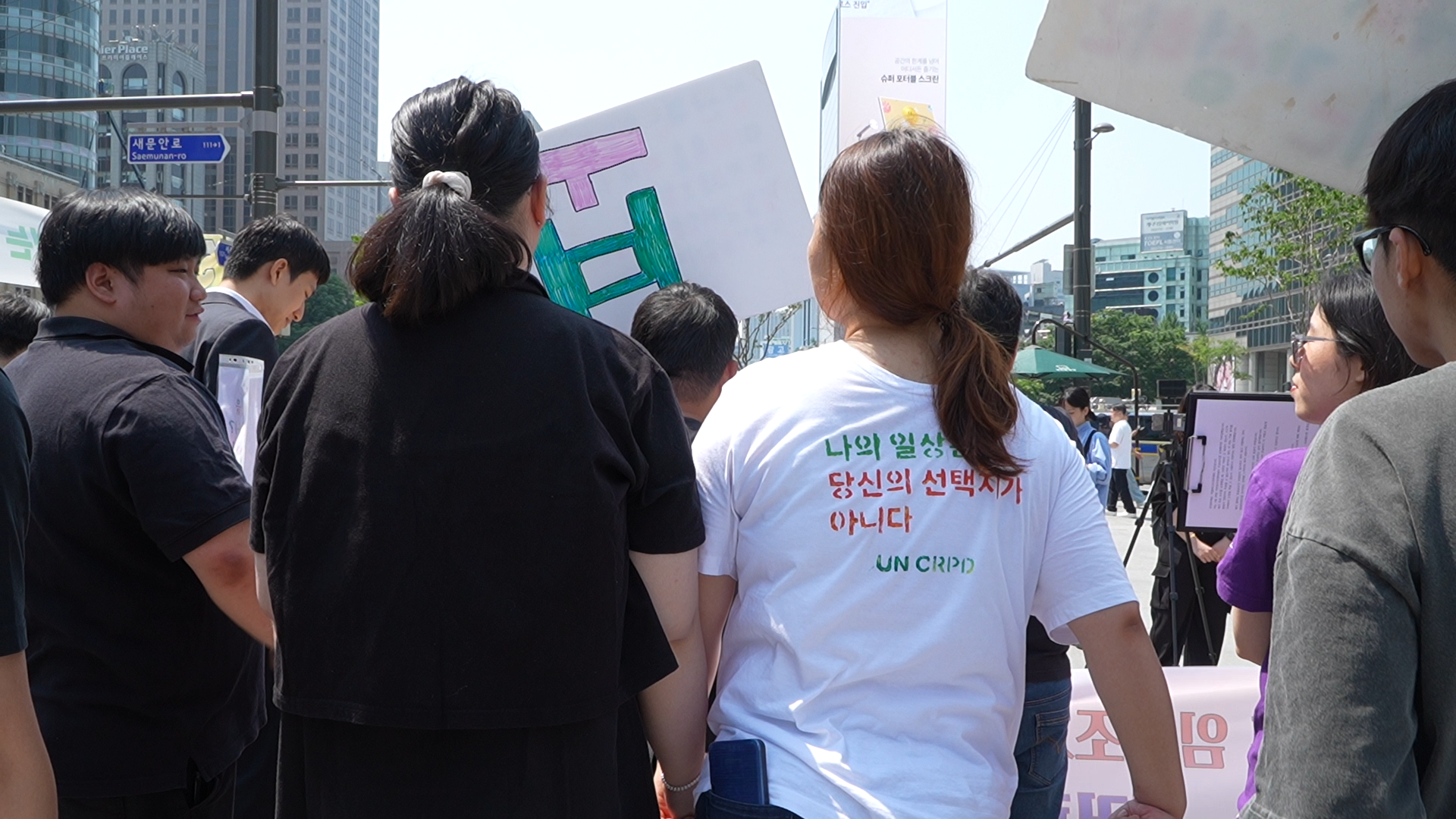 마스크를 쓴 두 사람이 앉아있다. 오른쪽 사람은 녹음용 마이크 앞에 앉아 헤드셋을 끼고 있고, 왼쪽 사람은 어딘가를 가리키고 있다.