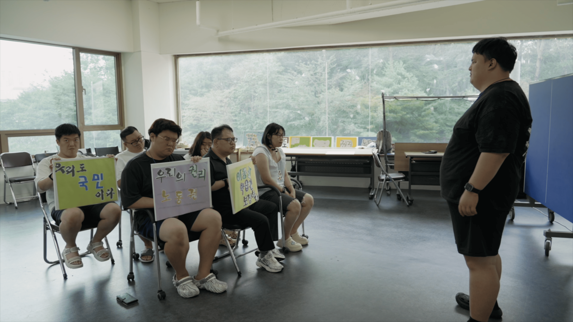 마스크를 쓴 두 사람이 앉아있다. 오른쪽 사람은 녹음용 마이크 앞에 앉아 헤드셋을 끼고 있고, 왼쪽 사람은 어딘가를 가리키고 있다.