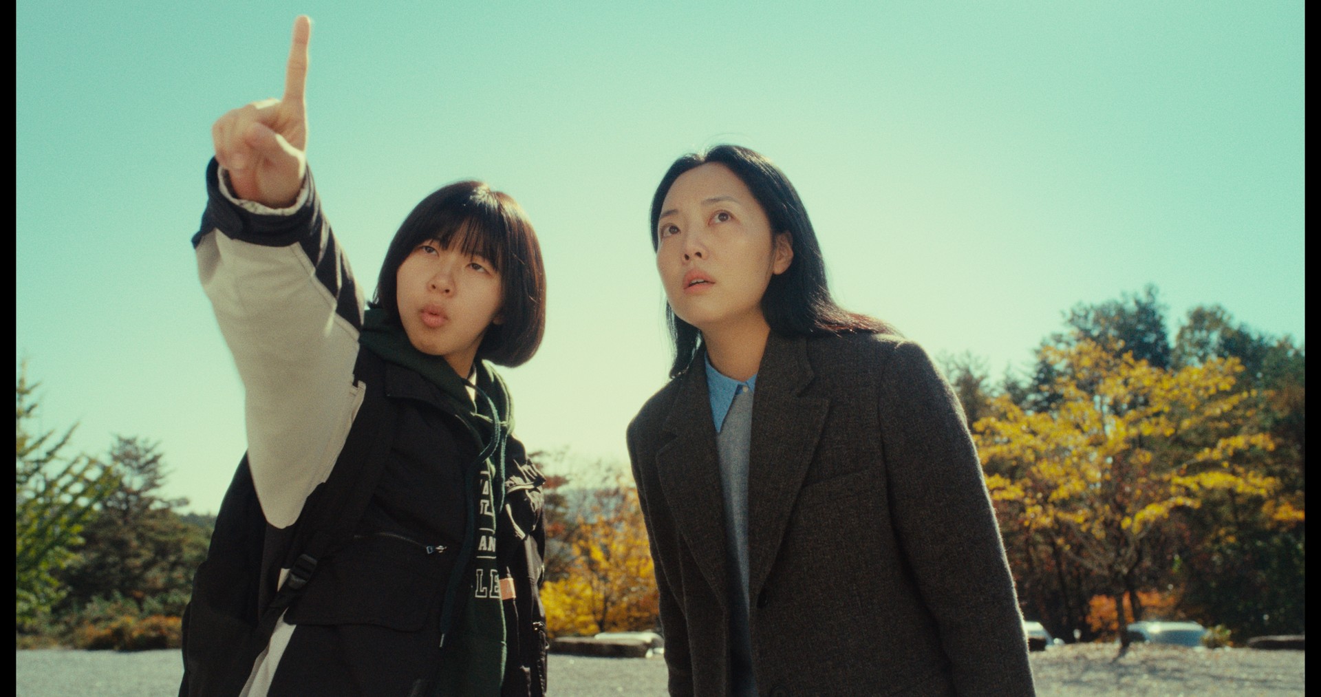 마스크를 쓴 두 사람이 앉아있다. 오른쪽 사람은 녹음용 마이크 앞에 앉아 헤드셋을 끼고 있고, 왼쪽 사람은 어딘가를 가리키고 있다.