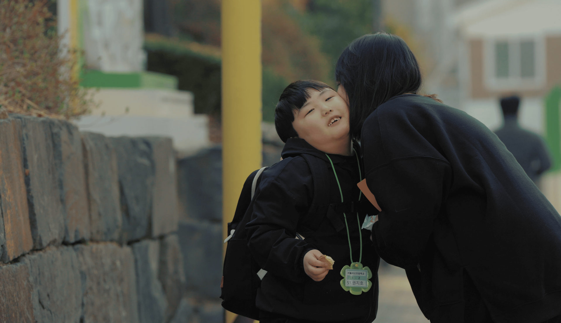 마스크를 쓴 두 사람이 앉아있다. 오른쪽 사람은 녹음용 마이크 앞에 앉아 헤드셋을 끼고 있고, 왼쪽 사람은 어딘가를 가리키고 있다.