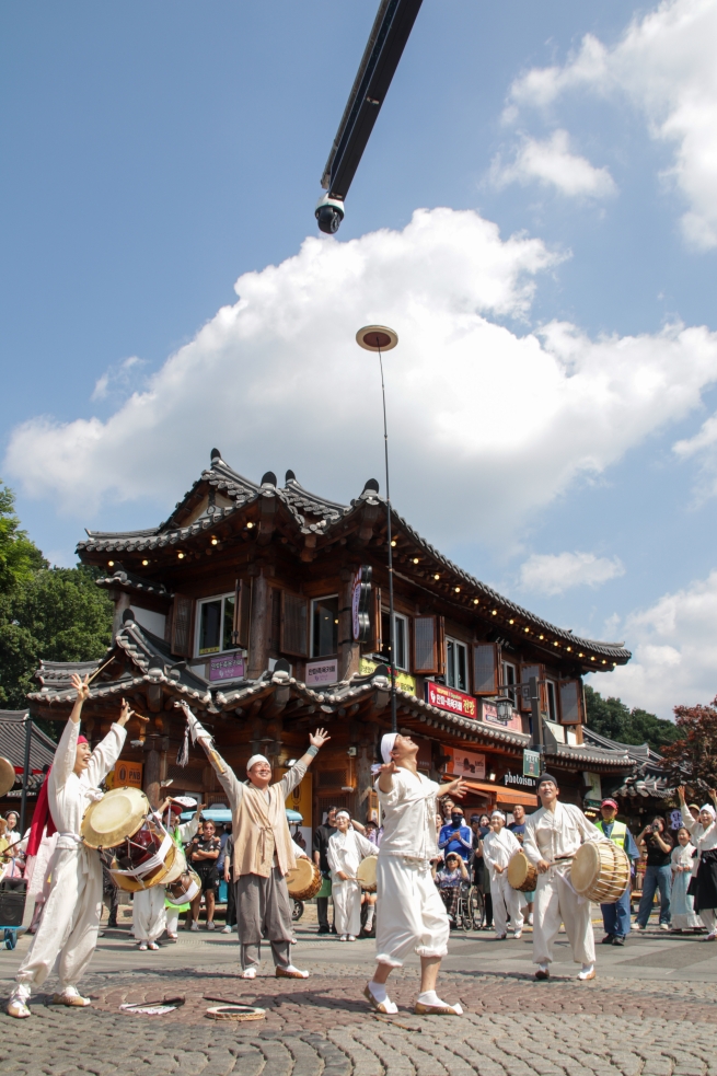 전주 한옥마을 거리에서 버나놀이 공연자가 5미터 길이의 긴 막대를 이마에 올려 균형을 잡으며 꼭대기에서 접시를 돌리는 장면. 전통 한옥 건물과 푸른 하늘을 배경으로 펼쳐지는 아슬아슬한 전통 기예 공연