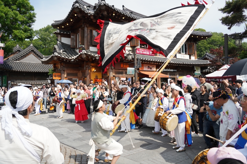 전주 한옥마을 전통연희 퍼레이드에서 전북무형문화재 전주기접놀이 용깃발을 공연자가 낮은 자세로 몸을 젖히며 역동적으로 휘두르는 장면. 풍물패 장단에 맞춰 한옥 건물과 맑은 하늘을 배경으로 수백 명의 관객들이 사진을 찍으며 감탄하고 있다