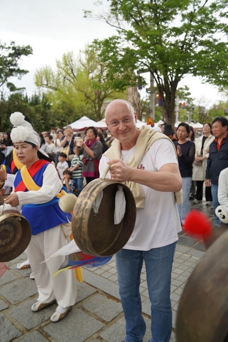 전주 한옥마을 전통연희 퍼레이드에서 서양인 남성 외국인 관광객이 풍물패 출연자 옆에서 징을 직접 치며 환하게 웃는 장면. 관객들이 지켜보는 가운데 풍물 장단에 함께 참여하고 있다
