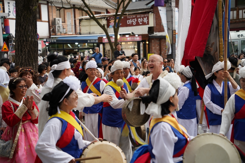 전주 한옥마을 전통연희 퍼레이드에서 서양인 외국인 관광객이 징을 직접 치며 풍물패 대열 한가운데에서 함께 악기 연주에 참여하는 장면. 북·장구 소리가 가득한 거리에서 한국인 연주자들과 외국인이 하나가 되어 풍물 장단을 이어가고 있다