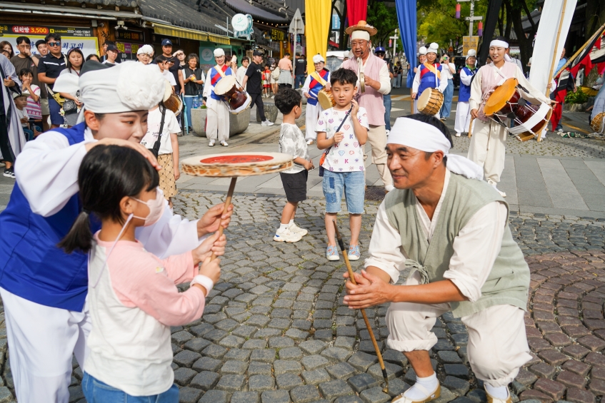 전주 한옥마을 전통연희 퍼레이드에서 어린 여자아이가 출연자의 도움을 받아 버나놀이 접시돌리기를 직접 체험하는 장면. 출연자가 아이의 눈높이에 맞춰 무릎을 굽히고 따뜻하게 이끌어 주고 있다