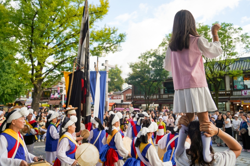 전주 한옥마을 전통연희 퍼레이드에서 어린 여자아이가 어른 어깨 위에 올라 주먹을 쥐고 풍물패를 향해 신나게 호응하는 장면. 아이의 시선으로 바라본 풍물패 행렬과 기접놀이 깃발이 펼쳐지는 생동감 넘치는 현장