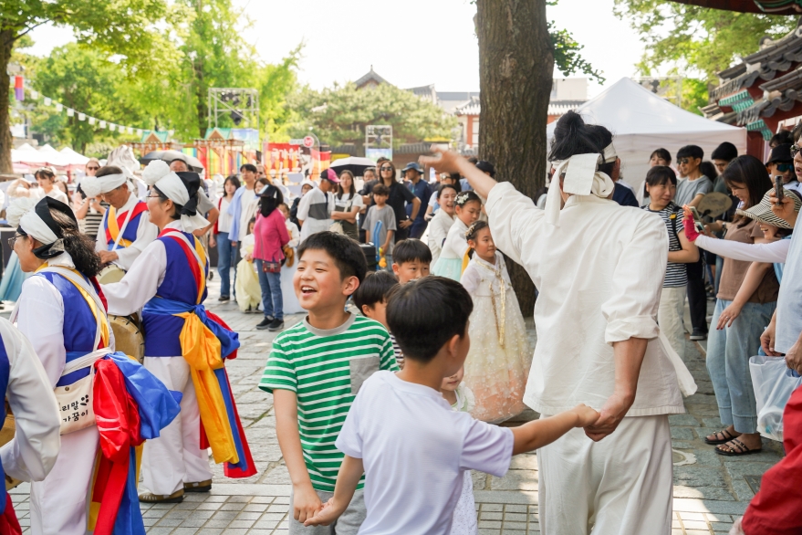전주 한옥마을 전통연희 퍼레이드 대동놀이 마당에서 어린이들이 출연자와 손을 잡고 함께 춤을 추며 활짝 웃는 장면. 경기전 앞 나무 그늘 아래 아이들과 가족 관객들이 어우러져 흥겨운 한때를 보내고 있다