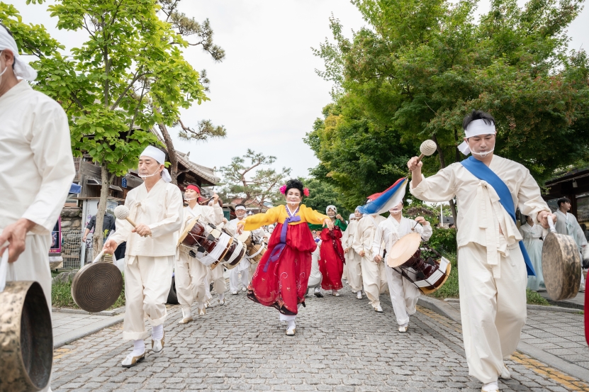 전주 한옥마을 전통연희 퍼레이드에서 화려한 빨간 한복 차림의 잡색광대가 풍물패 행렬 한가운데서 흥겹게 춤을 추며 앞으로 나아가는 장면. 소나무와 한옥 처마를 배경으로 돌바닥 거리에 펼쳐지는 전통 행렬의 생동감이 느껴진다