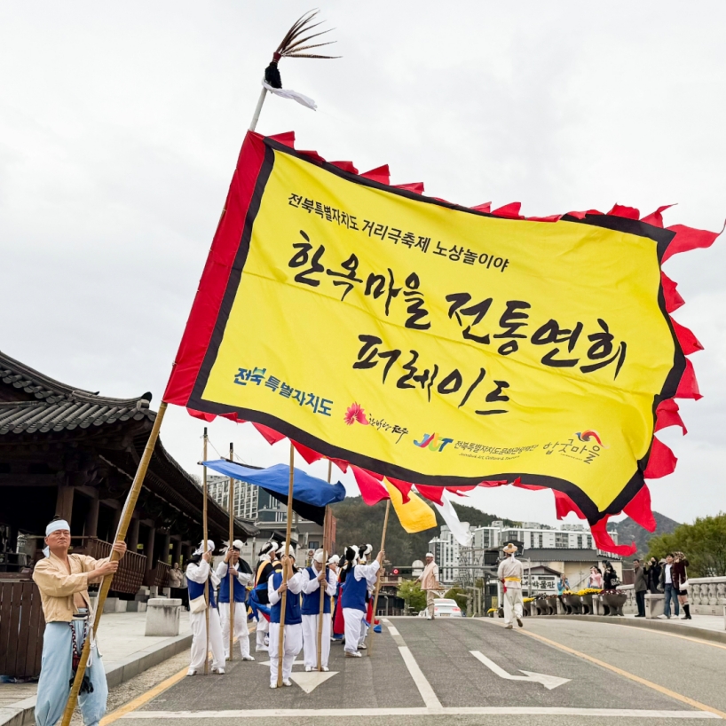 전주 한옥마을 전통연희 퍼레이드 시작 지점인 남천교에서 '한옥마을 전통연희 퍼레이드' 공식 현수기를 든 공연자가 풍물패 행렬을 이끌며 출발하는 장면. 전북특별자치도·전주시·합굿마을 로고가 선명하게 새겨진 노란 깃발이 하늘을 향해 펼쳐지고 있다