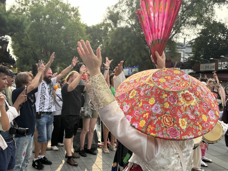전주 한옥마을 전통연희 퍼레이드에서 화려한 꽃무늬 모자를 쓴 출연자가 부채를 들고 외국인 관광객들과 함께 전주 구전 노동요 떼창을 이끄는 장면. 다국적 외국인 관객들이 손을 들고 함께 호응하고 있다