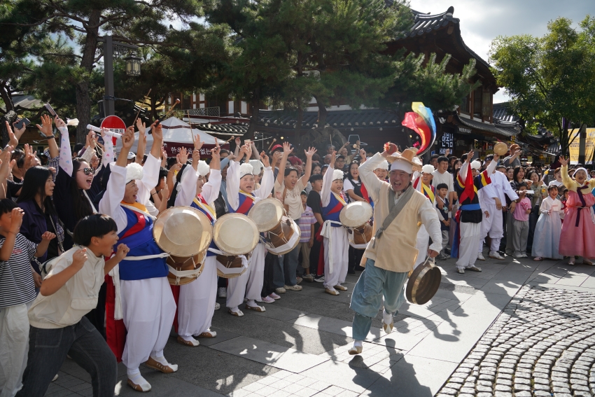 전주 한옥마을 경기전 앞에서 풍물패 출연자가 수백 명의 관객들과 함께 전주 구전 노동요 떼창을 이끄는 장면. 관객들이 두 손을 들고 함께 노래하며 130년 전 전주의 민속 문화를 체험하고 있다