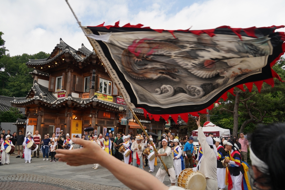 전주 한옥마을 거리에서 전북무형문화재 전주기접놀이 용깃발이 전통 한옥 건물을 배경으로 크게 펼쳐지는 장면. 풍물패가 북과 장구를 치며 행진하고, 관객들이 행렬 주위로 모여 함께하고 있다
