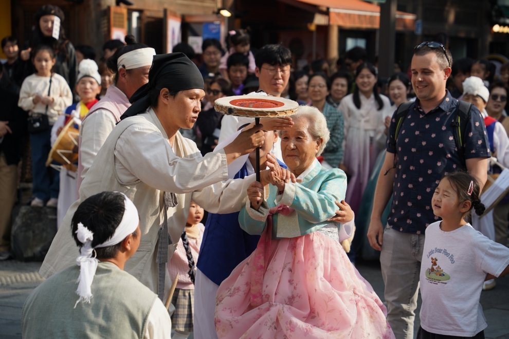 전주 한옥마을 전통연희 퍼레이드에서 한복 차림의 어르신 할머니가 출연자의 도움을 받아 버나놀이 접시돌리기를 직접 체험하는 장면. 외국인 관광객과 어린이를 포함한 관객들이 따뜻한 미소로 지켜보고 있다