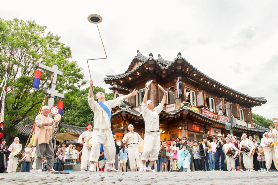 전주 한옥마을 거리에서 전통 한복 차림의 공연자들이 버나놀이(접시돌리기)를 선보이는 장면. 배경에는 전통 한옥 건물과 기접놀이 깃발이 보이고, 많은 관객들이 공연을 둘러싸고 지켜보고 있다