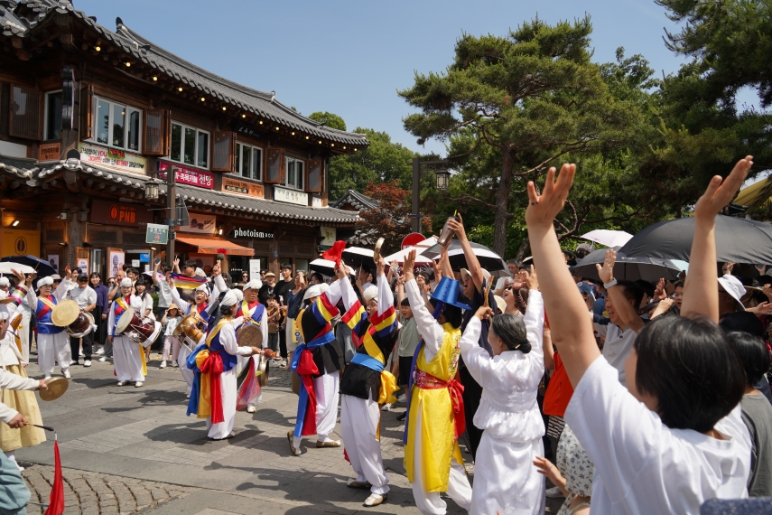전주 한옥마을 거리에서 풍물패 출연자들과 관객들이 함께 손을 들고 전주 구전 노동요 떼창에 참여하는 장면
