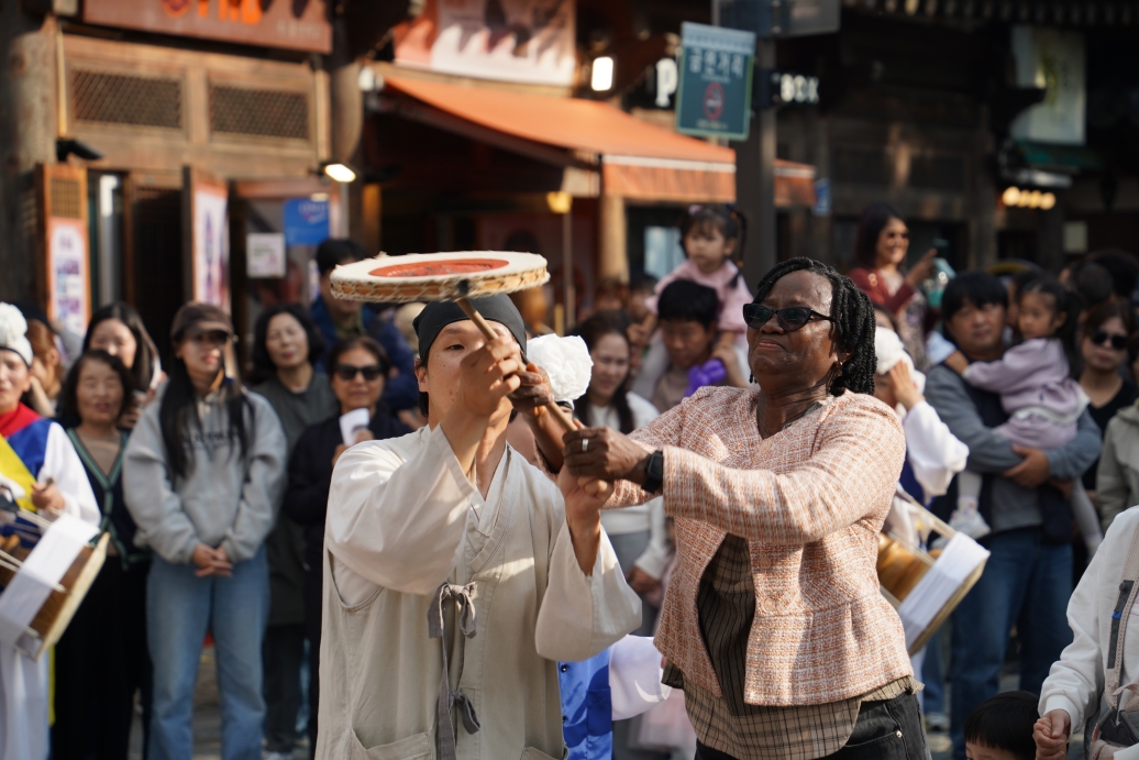 전주 한옥마을 전통연희 퍼레이드에서 외국인 관광객이 출연자와 함께 버나놀이 접시돌리기를 직접 체험하는 장면. 두 사람이 함께 막대를 잡고 접시를 올려 돌리며 교감하고 있고, 주변 관객들이 미소 지으며 지켜보고 있다