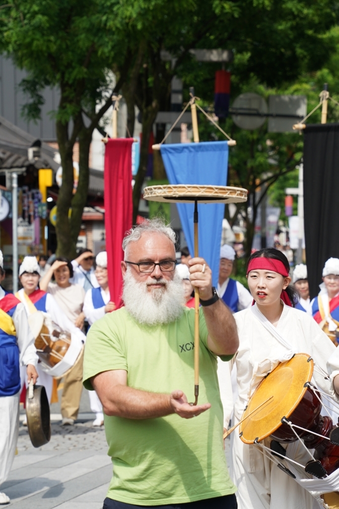 전주 한옥마을 전통연희 퍼레이드에서 흰 수염의 서양인 외국인 관광객이 버나놀이 막대를 직접 들고 풍물패 행렬 맨 앞에서 함께 걸어가는 장면. 출연자와 어깨를 나란히 하며 퍼레이드에 완전히 참여하고 있다