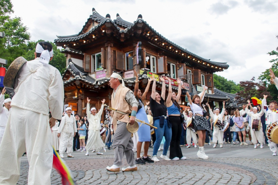 전주 한옥마을 전통연희 퍼레이드에서 외국인 관광객들이 직접 무대 앞에 나와 전주 구전 노동요 떼창을 손을 높이 들며 함께 지휘해보는 장면. 전통 한옥 건물을 배경으로 한국인과 외국인이 하나가 되어 130년 전 전주의 노래를 함께 부르고 있다
