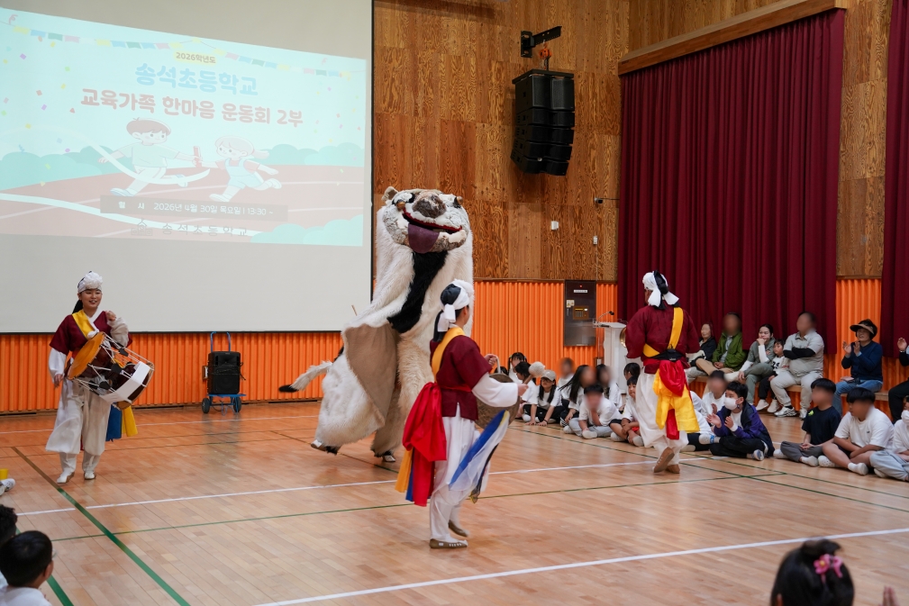 풍물단의 장구와 북 연주에 맞춰 강아지 탈 캐릭터 해피가 머리를 높이 올리며 사자탈춤 공연을 펼치고 있다. 대형 스크린에 송석초등학교 한마음 운동회 영상이 투사되고있다