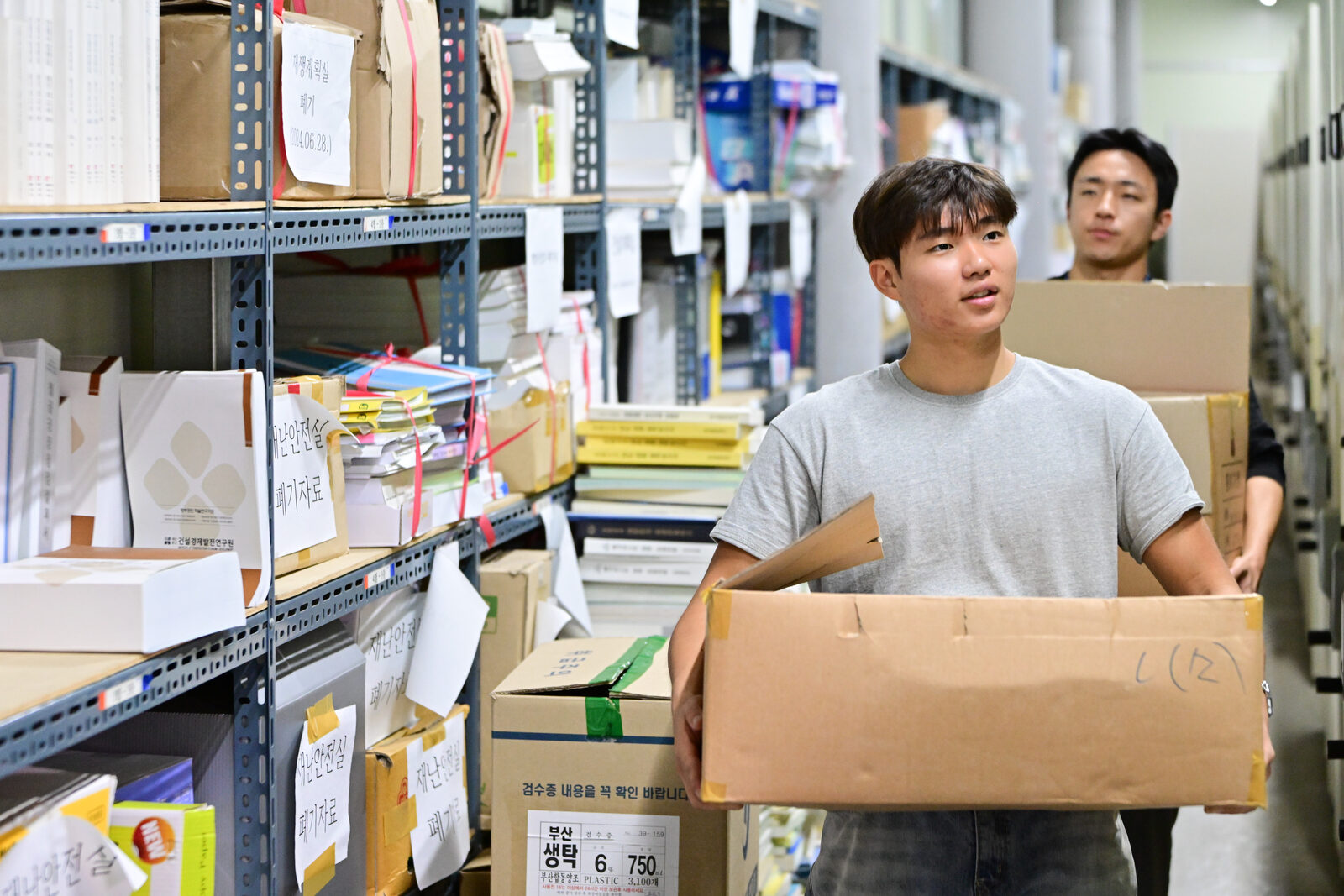 회색 반팔을 입은 오혜성 사원이 종이상자를 안고 기록관 통로를 걸어 나오고, 뒤에서 검정 긴팔을 입은 정우영 사원이 같은 상자를 들고 따라오는 모습