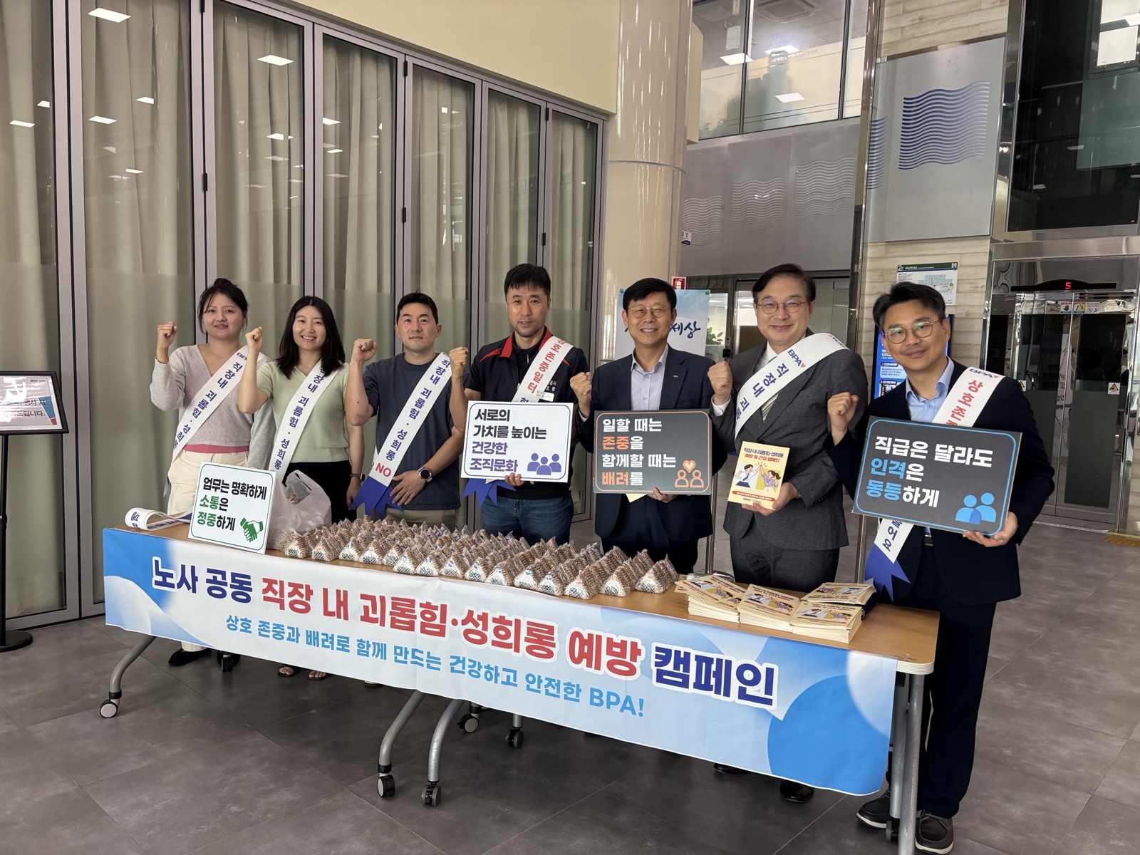 사옥 로비에 「노사 공동 직장 내 괴롭힘·성희롱 예방 캠페인」 현수막을 두른 배부대가 놓여 있고, 어깨띠를 맨 노사 대표와 직원 일곱 명이 ‘서로의 가치를 높이는 건강한 조직문화’, ‘일할 때는 존중을 함께할 때는 배려를’, ‘직급은 달라도 인격은 동등하게’ 같은 문구가 적힌 손팻말을 들고 서 있다. 탁자에는 나눠줄 간식과 예방 리플릿이 쌓여 있다