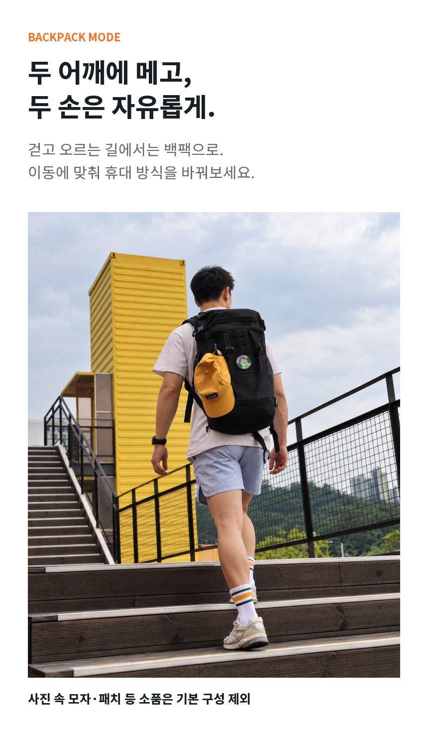 남성 모델이 후크건을 세로 백팩 형태로 메고 야외 계단을 오르는 착용 이미지. 모자와 패치는 기본 구성 제외.