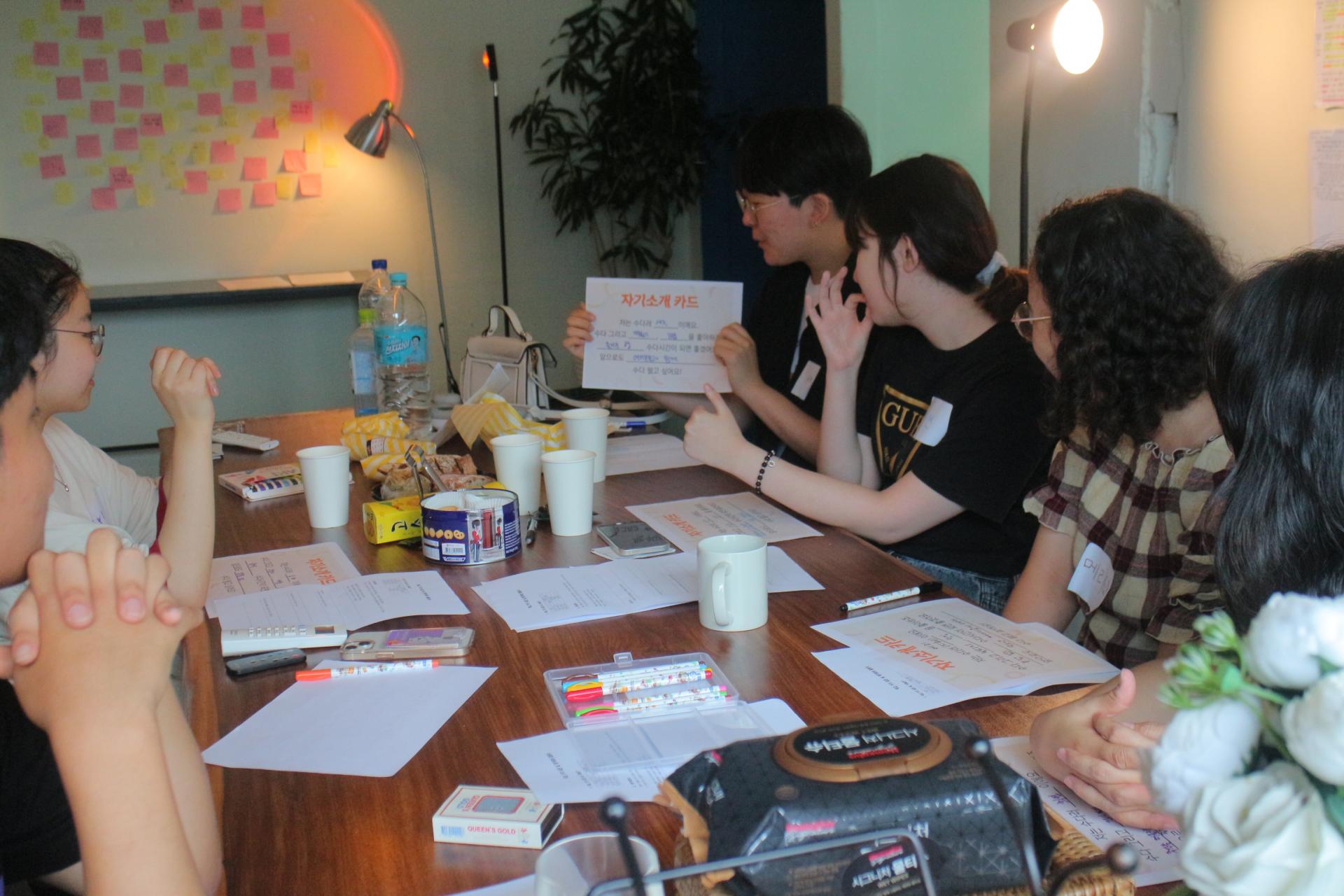 참여자들이 책상에 모여 앉아있는 사진. '자기소개 카드'라고 적힌 종이를 들고 발표하는 참여자를 보고 있다.