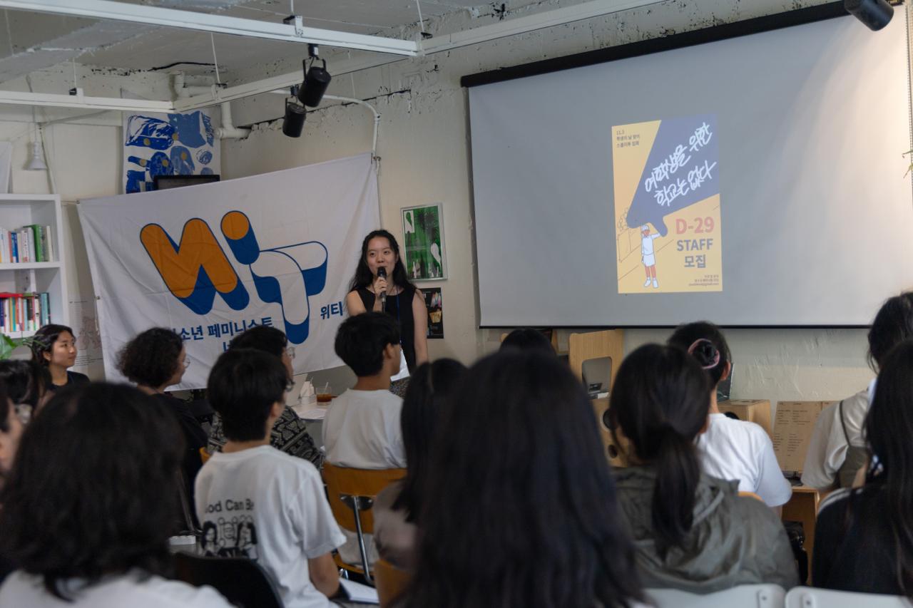 활동가 경하가 마이크를 잡고 말하고 있다. 스크린에는 '여학생을 위한 학교는 없다' 포스터가 떠있다. 