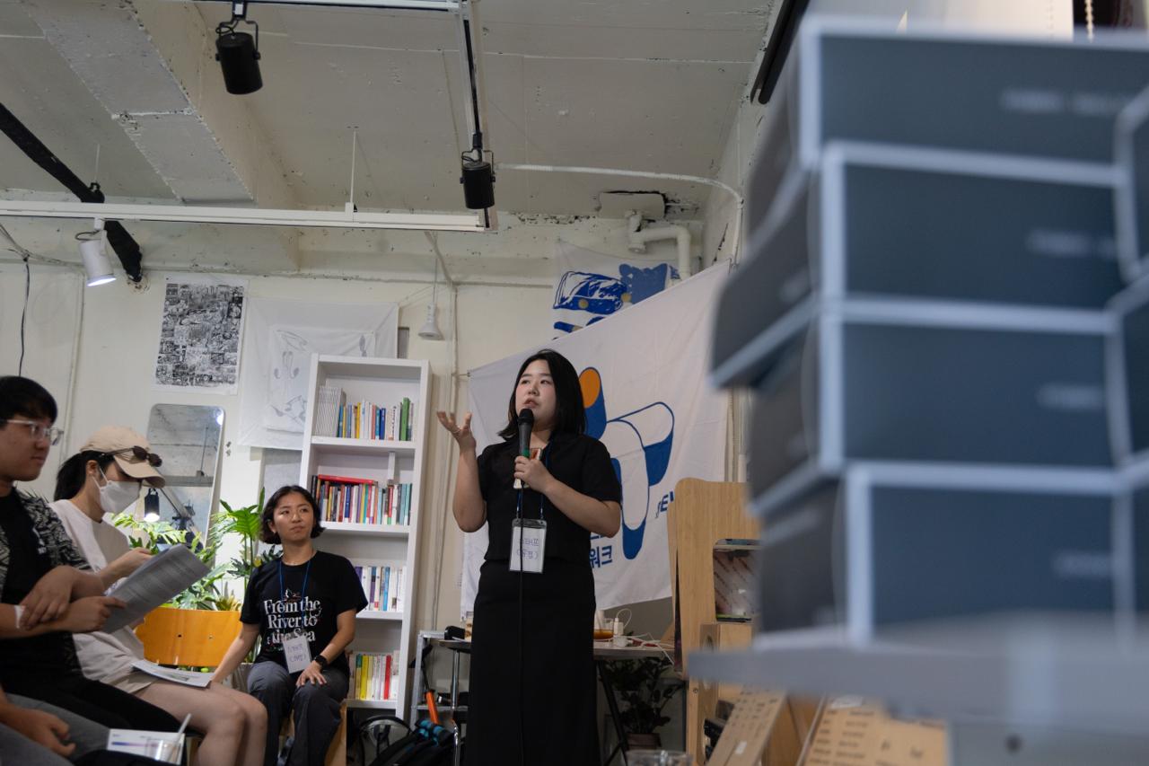 활동가 유경이 마이크를 잡고 말하고 있다. 참가자들이 진지한 얼굴로 듣고 있다. 