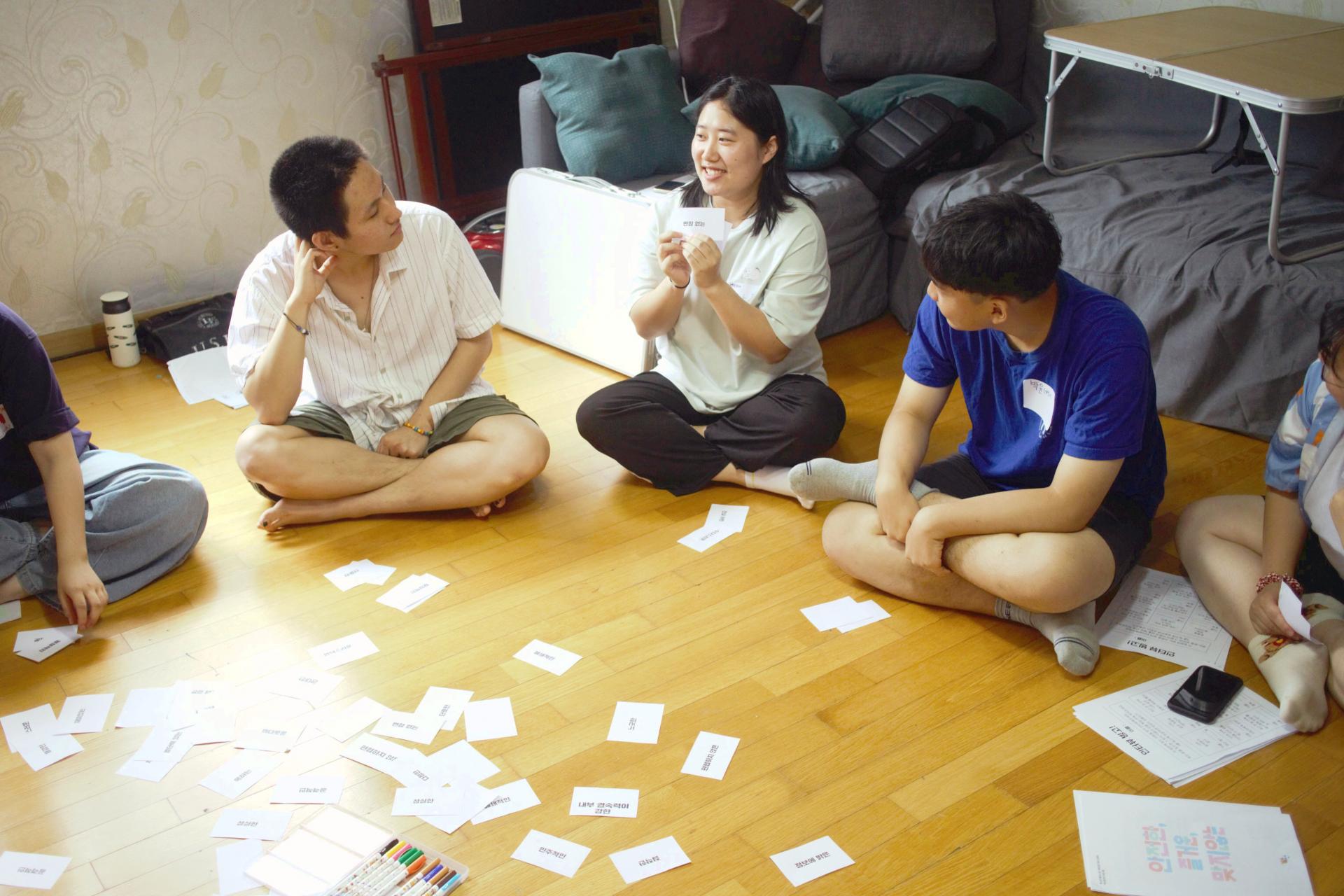 참가자들이 거실 바닥에 둘러 앉아있다. 중앙에는 글자들이 적힌 작은 카드들이 널려있다.