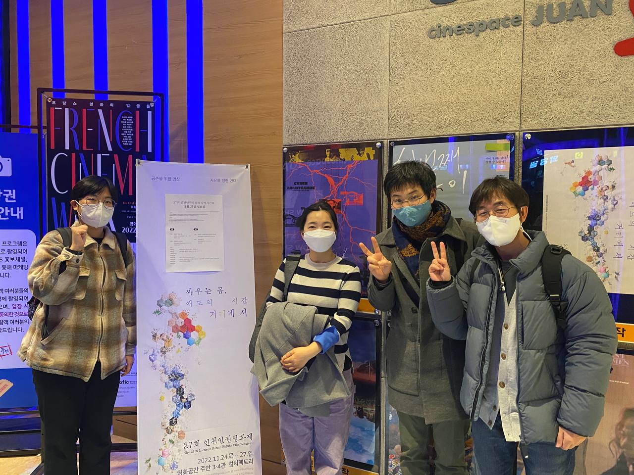 "싸우는 몸, 애도의 시간 거리에서 27회 인천인권영화제"라고 써져있는 세로형 배너 왼쪽에 한 명이 주먹을 쥐고 카메라를 보고 서있다. 배너 오른편으로 세 명의 사람이 카메라를 보고 서있다. 가장 오른쪽 두 사람은 손가락 두개로 '브이'표시를 하고 있다.