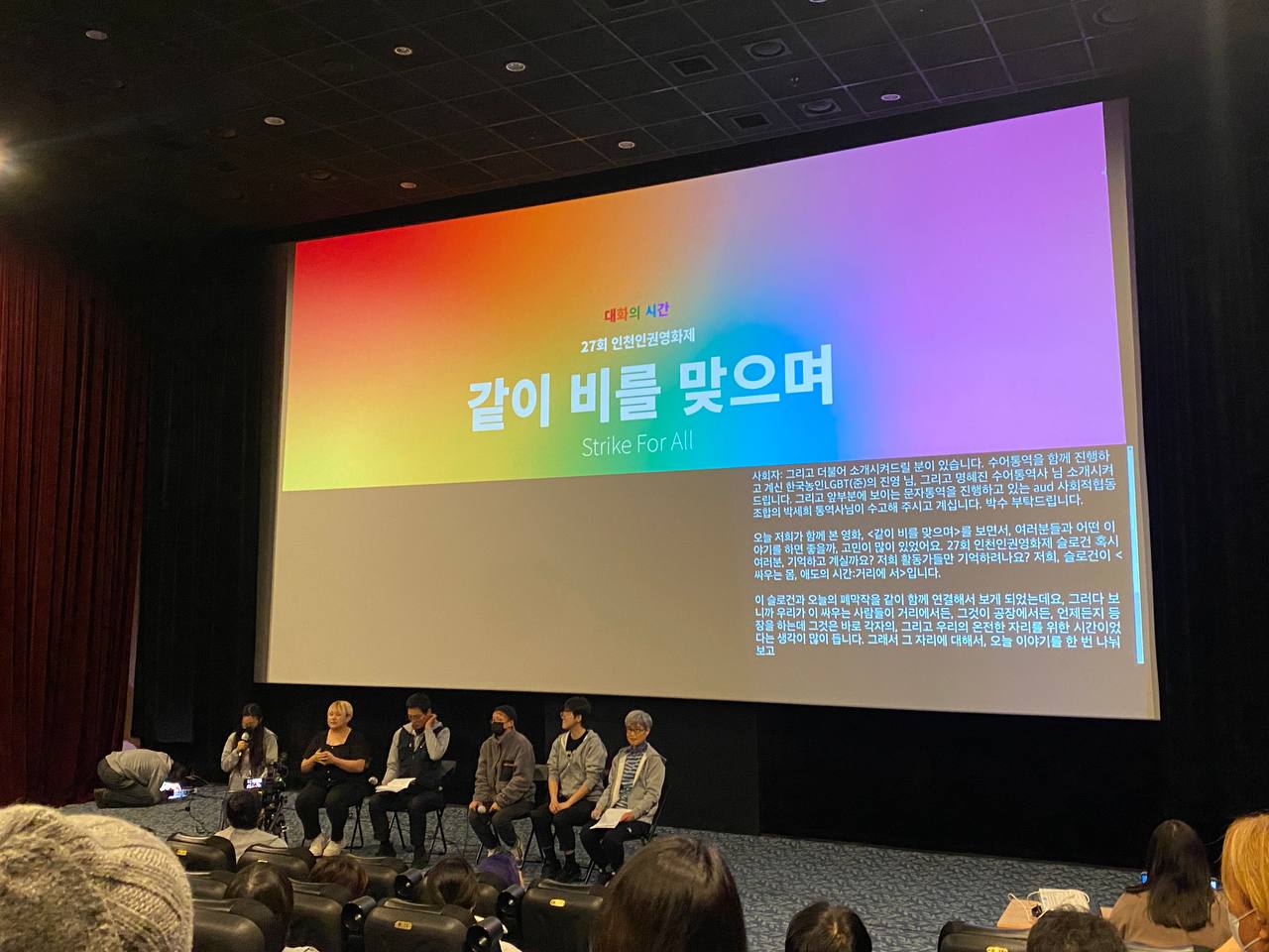 영화관 사진. 영화관 앞쪽 스크린에 무지개 빛 배경 위로 "대화의 시간 27회 인천인권영화제 같이 비를 맞으며 Strike For All" 이라는 하얀 자막이 쓰여있고 오른쪽 아래로 수어통역 타이핑 글씨가 보인다.  스크린 앞으로 <대화의 시간> 순서에 참여하는 사회자와 패널들이 총 여섯 명 앉아있다.