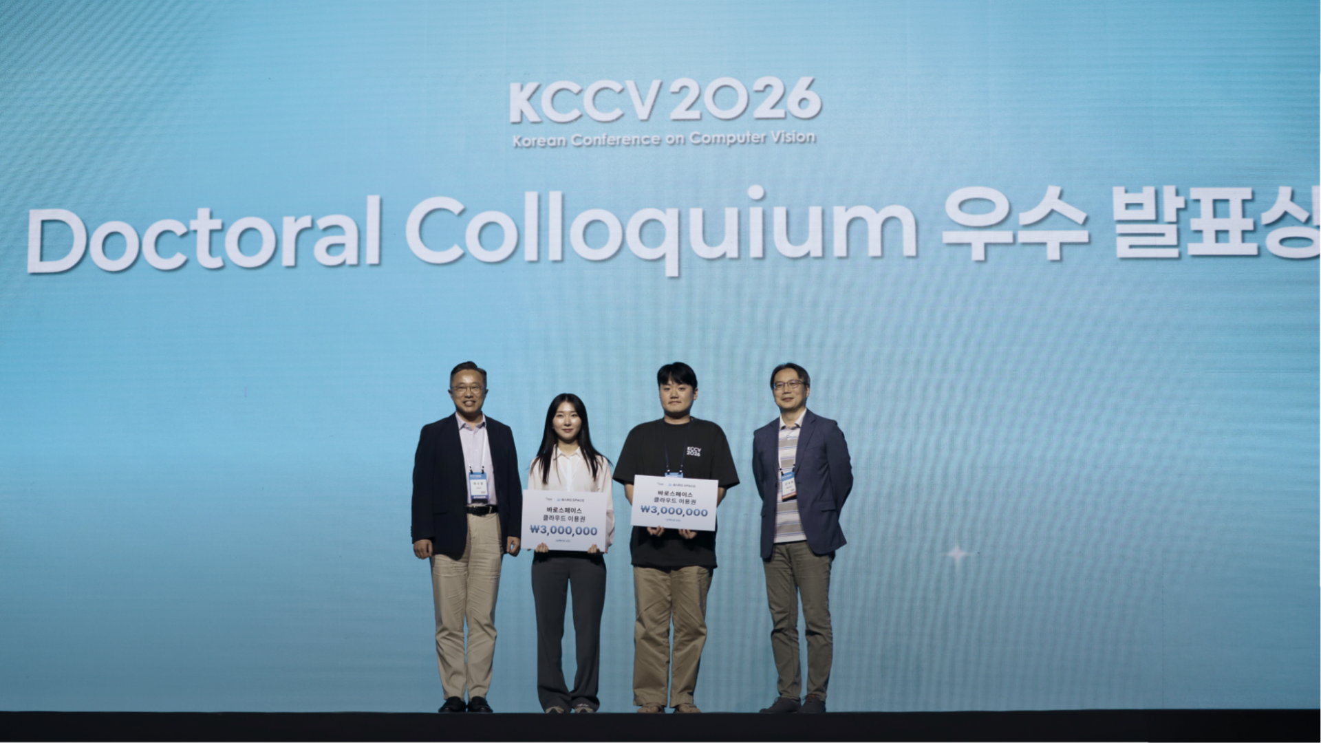 KCCV 2026 Doctoral Colloquium 우수 발표상 시상식 무대에서 수상자 2인이 각 300만원 상당의 바로스페이스(BARO SPACE) 클라우드 이용권을 들고 관계자들과 기념 촬영하는 모습을 다루는 바로AI(BARO AI) 테크 블로그 자료입니다.