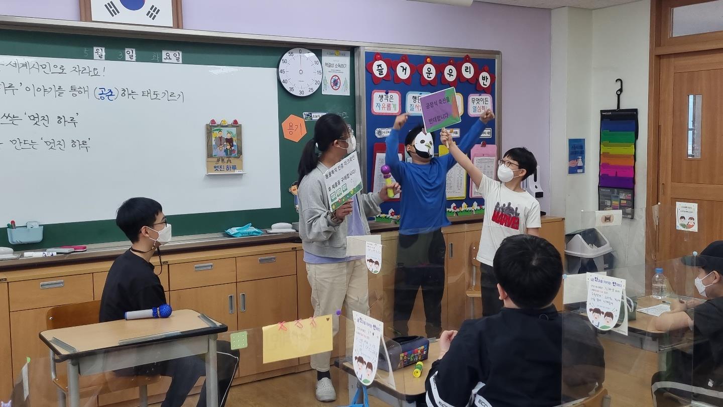 세 명의 학생이 칠판 앞으로 나와 역할극을 하고 있다. 한 명은 가면을 쓰고, 두 명은 동물복지 인증 마크 확인 후 제품을 구매합시다, 공장식 축산을 반대합니다라는 피켓을 들고 있다.