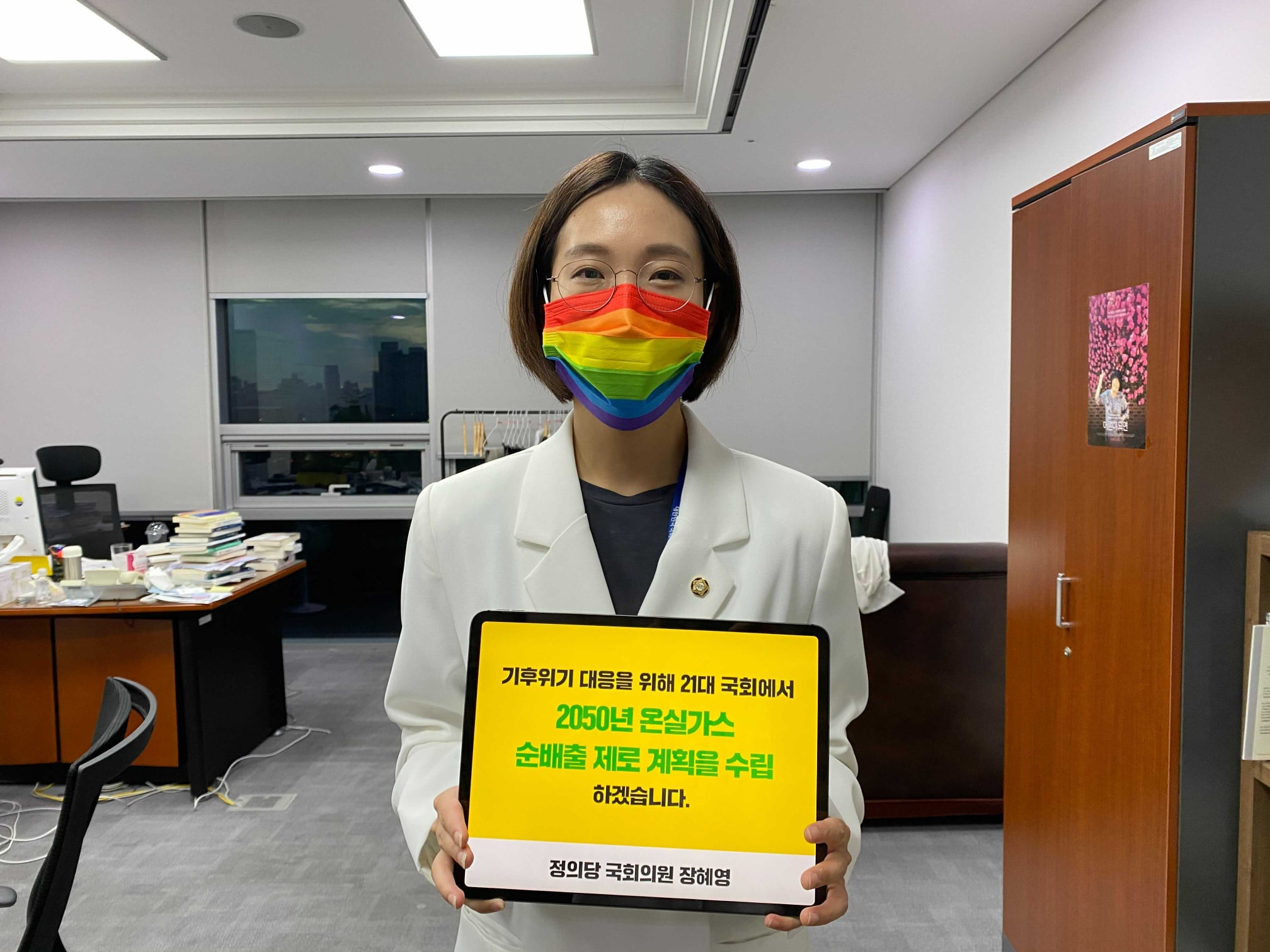 장혜영 의원이 국회의원회관 의원실에서 "기후위기 대응을 위해 21대 국회에서 2050년 온실가스 순배출 제로 계획을 수립하겠습니다" 문구가 적힌 태블릿pc를 들고, 무지개색 마스크를 착용한 모습