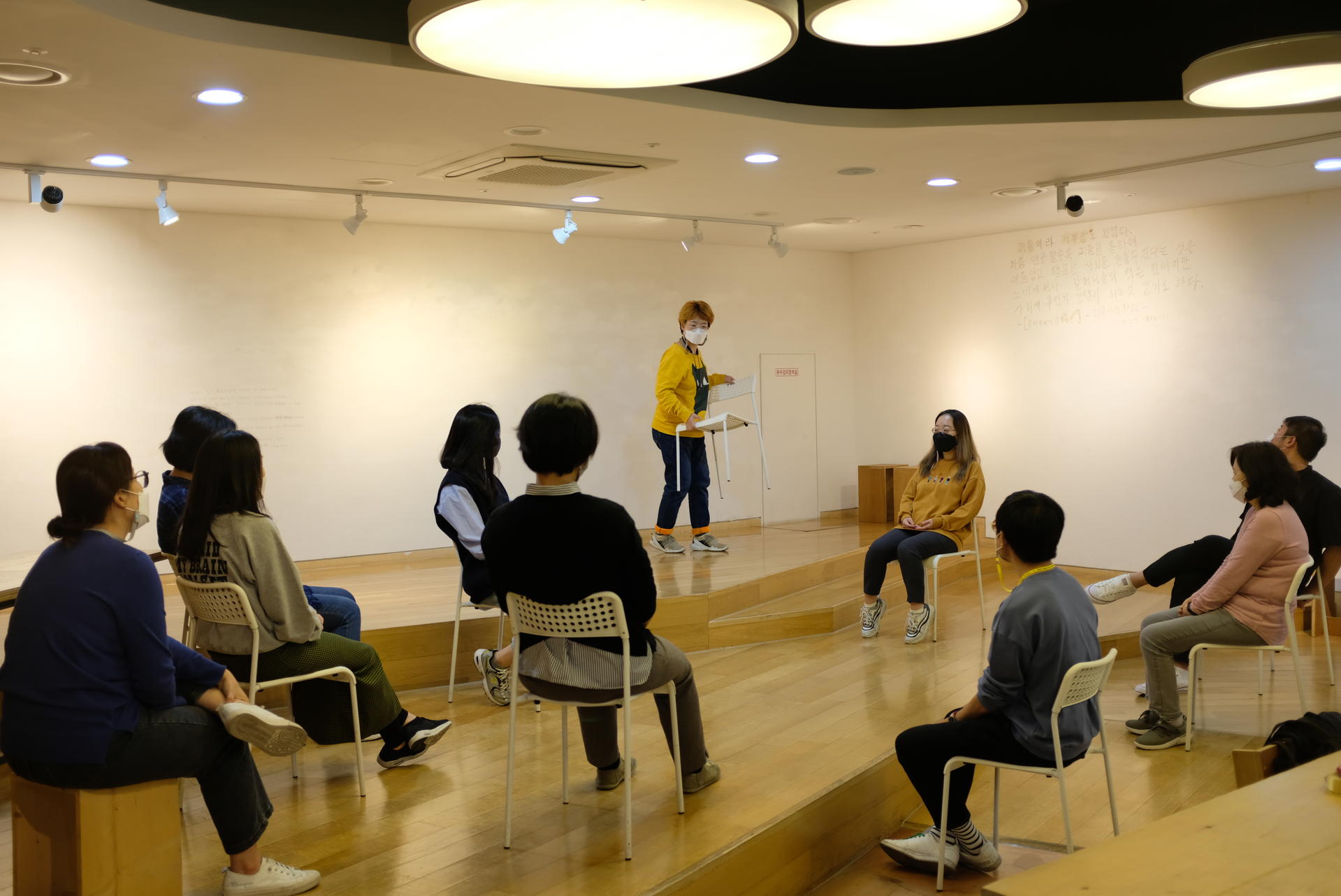 한 참가자가 의자를 들고 편안한 자리를 찾아 움직이고 있다. 다른 참가자들이 이 모습을 지켜보고 있다.