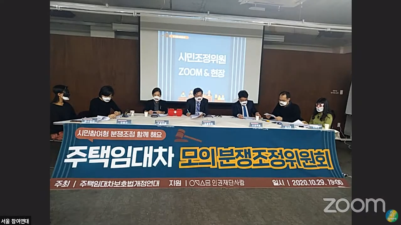 주택임대차 모의분쟁조정위원회가 적힌 현수막 뒤로 테이블이 있다. 테이블에는 7명이 앉아서 모의분쟁을 진행하고 있다.