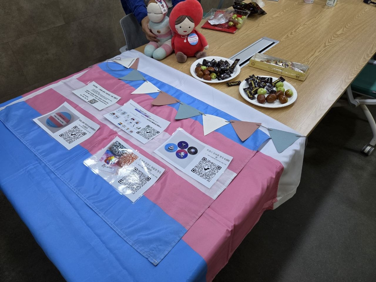 TGG 현장 사진. 트랜스젠더 프라이드 깃발 위에 후원 굿즈 안내가 보이고, 그 너머로 상에 다과와 발언 인형들이 보인다.