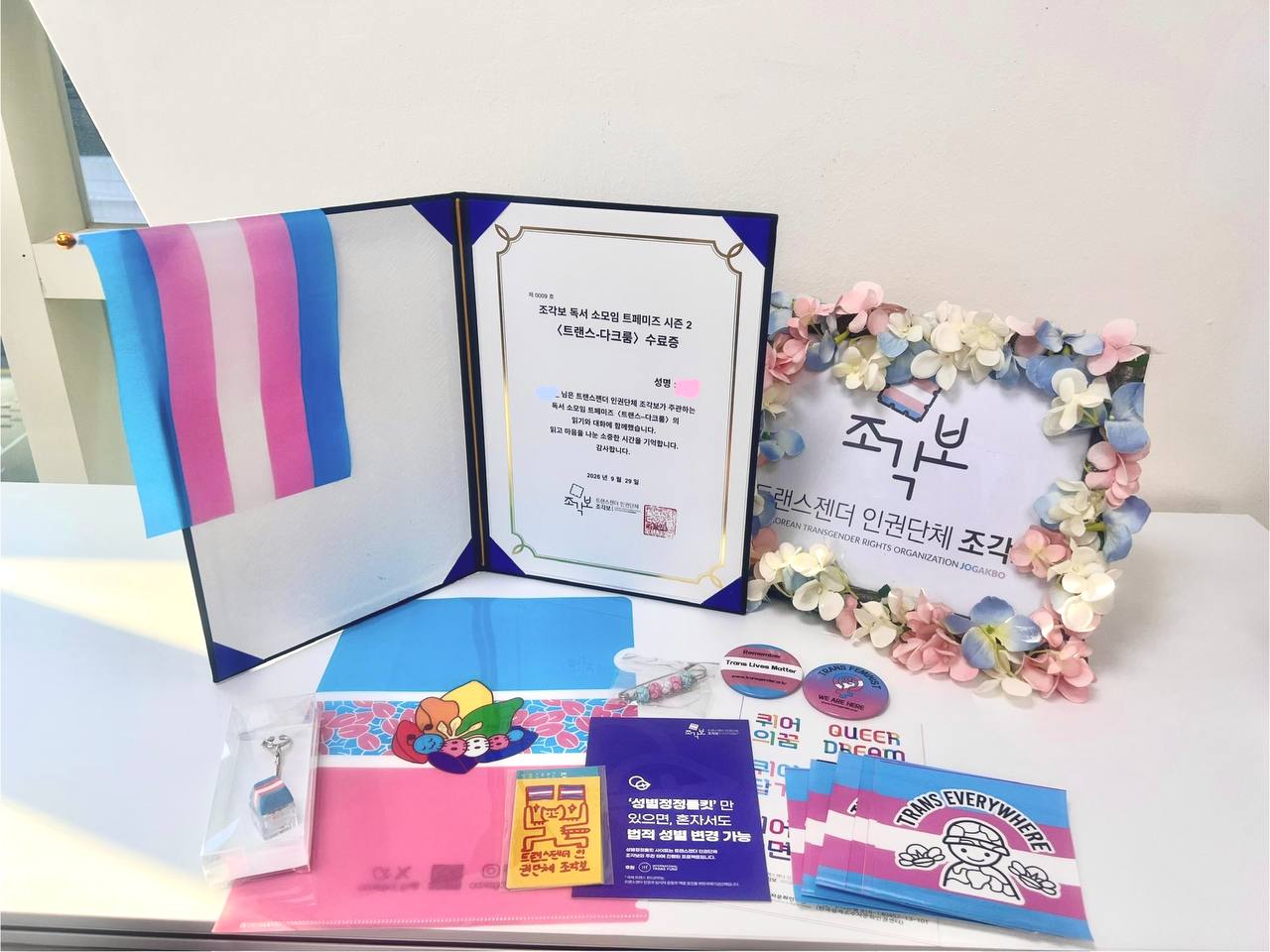 트랜스젠더 인권단체 조각보의 명패와 트페미즈 시즌 2 수료증, 굿즈들의 사진. 굿즈로는 트랜스 플래그 손깃발, 키링, L파일, 각종 스티커, 매듭옷핀, 핀버튼 뱃지, 그리고 성별정정툴킷 안내문이 보인다.