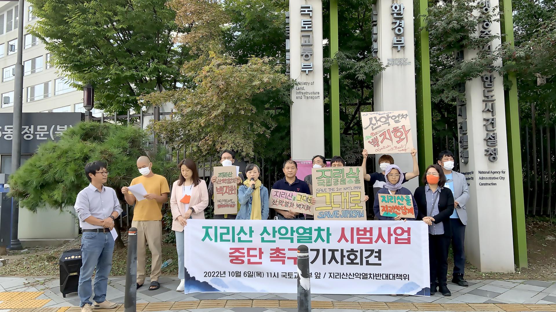 기자회견에 참석한 10여명의 시민과 '지리산 산악열차 시범사업 중단 촉구 기자회견' 현수막을 보이게 들고 있다. '산악열차 백지화', '지리산을 지리산답게', '지리산 국립공원을 그대로', '지리산을 그대로 산악열차 백지화' 등의 파켓을 들고 있는 시민들이 서있다. 뒤로는 국토교통부와 환경부 간판이 보인다.
