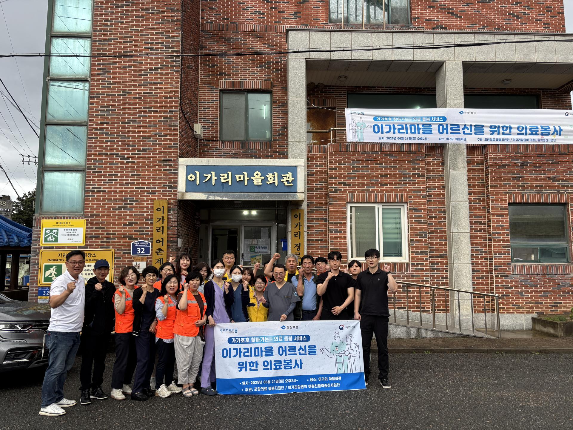 포항 이가리마을회관 앞에서 열린 의료봉사 행사. 의료진과 자원봉사자들이 마을 어르신들을 위해 진행된 ‘찾아가는 행복병원’ 의료봉사 활동 후 단체 기념촬영을 하고 있다. 건물에는 ‘이가리마을 어르신을 위한 의료봉사’ 현수막이 걸려 있다.