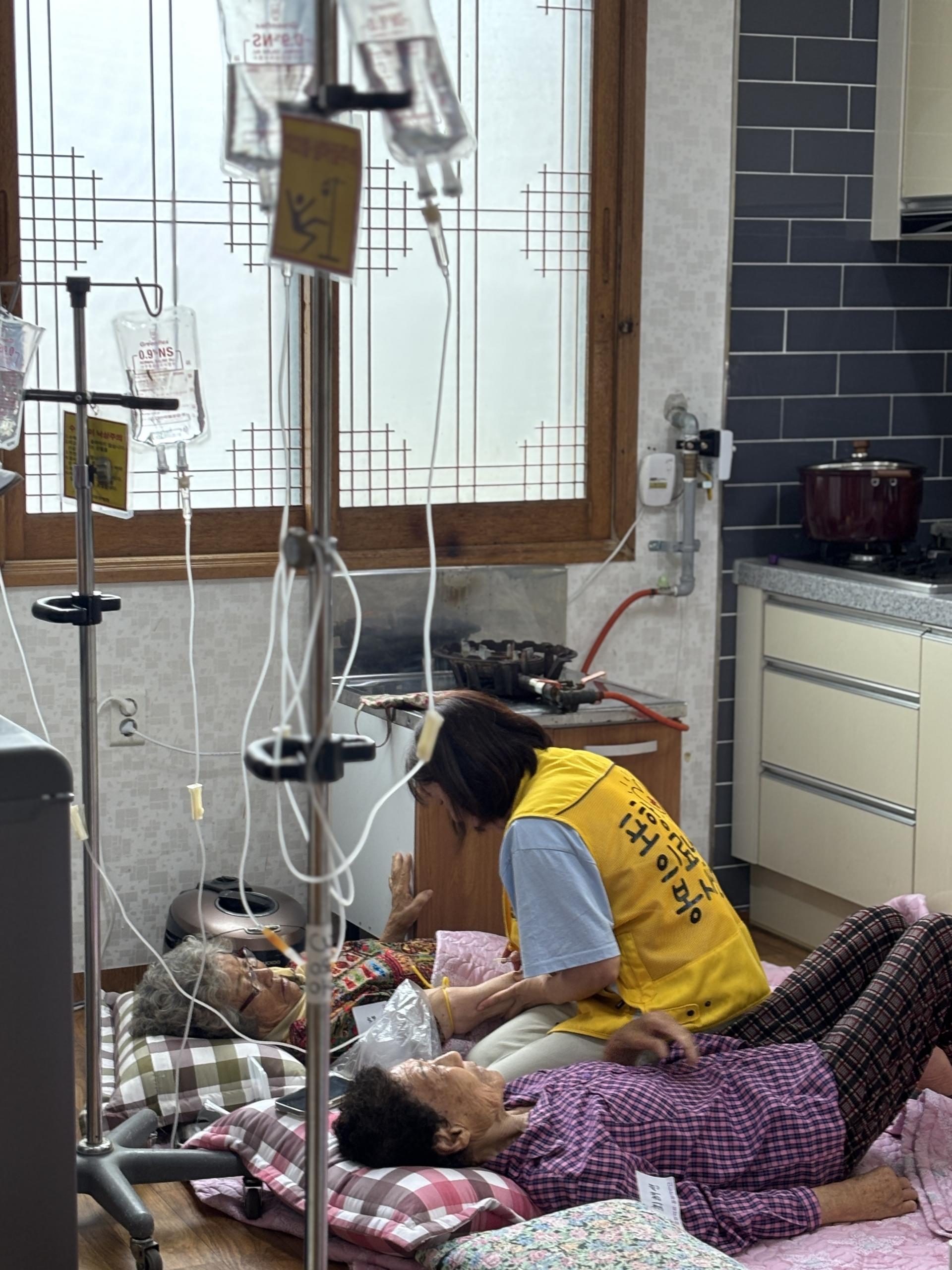 "이가리마을 의료봉사 현장. 노란 조끼를 입은 자원봉사자가 바닥에 누운 두 명의 할머니 곁에서 손을 잡고 대화를 나누며 돌보고 있다. 주변에는 링거대와 의료 장비가 설치되어 있으며, 따뜻한 분위기 속에서 어르신들을 보살피는 모습이 담겨 있다.