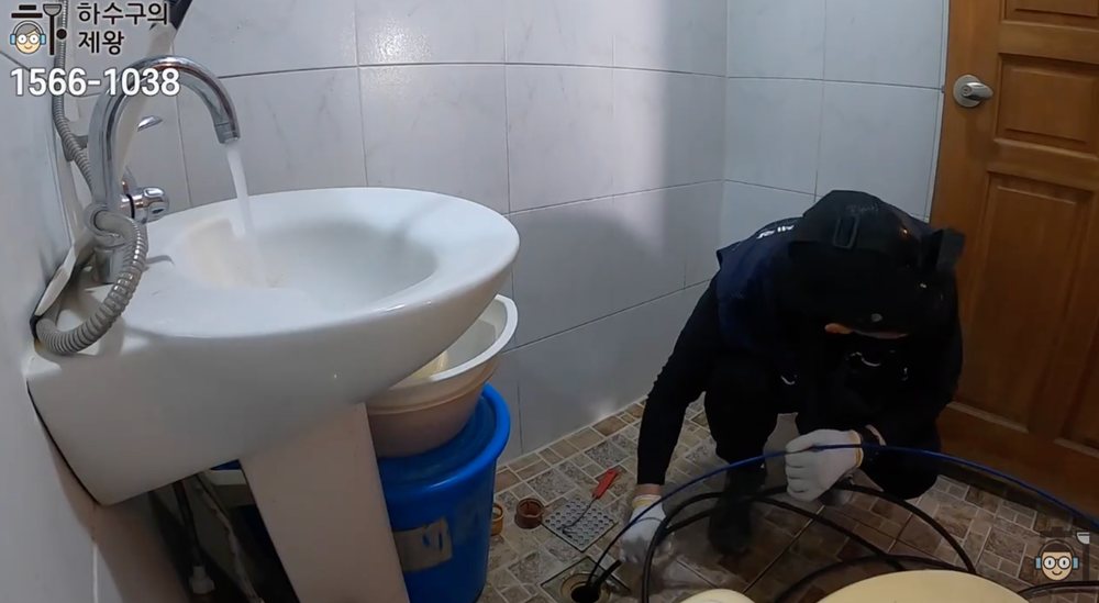 작업자가 화장실 바닥 배수구에 내시경과 청소 장비를 넣어 배관 내부를 점검하고 있는 장면