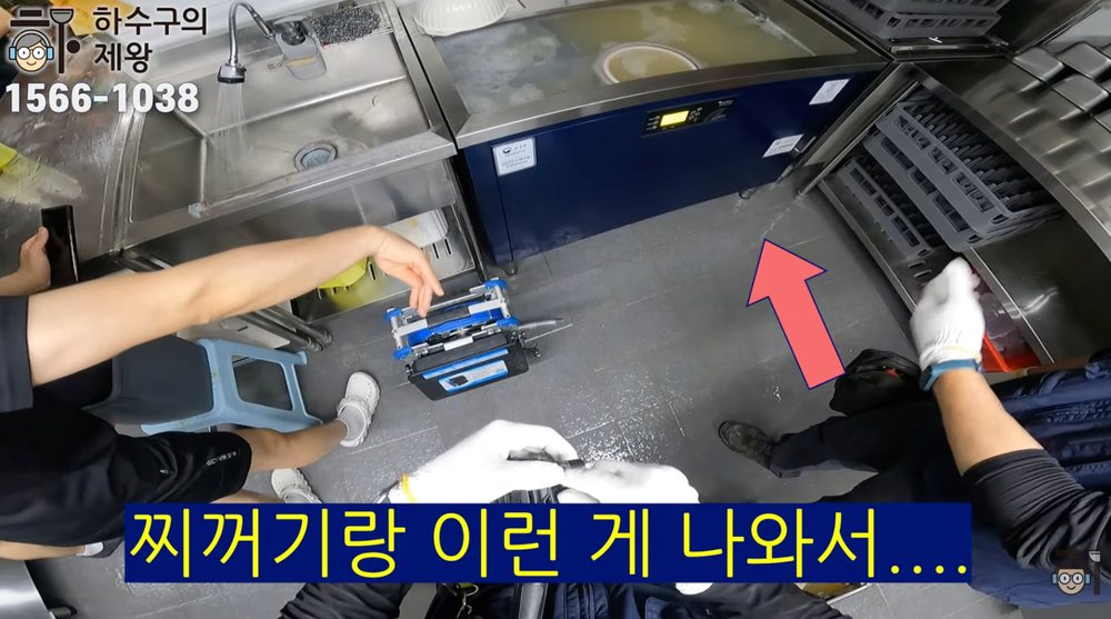 식당 싱크대 주변 바닥에 하수구 물이 넘쳐 고여 있고 작업자가 역류 위치를 확인하는 장면