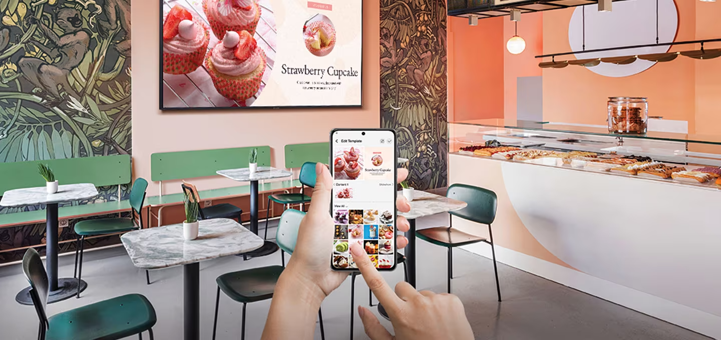 매장 안 벽에 제품이 설치되어 있고 딸기 케익 화면이 나오고 있습니다. 사용자가 모바일 비즈니스 TV 앱을 사용하고 있습니다.