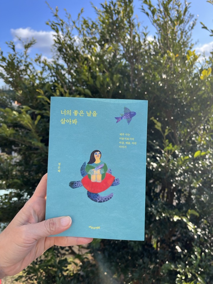 '너의 좋은 날을 살아봐' 책을 한손으로 들고 있다 뒤에는 초록잎이 무성한 나무와 하늘이 보인다.