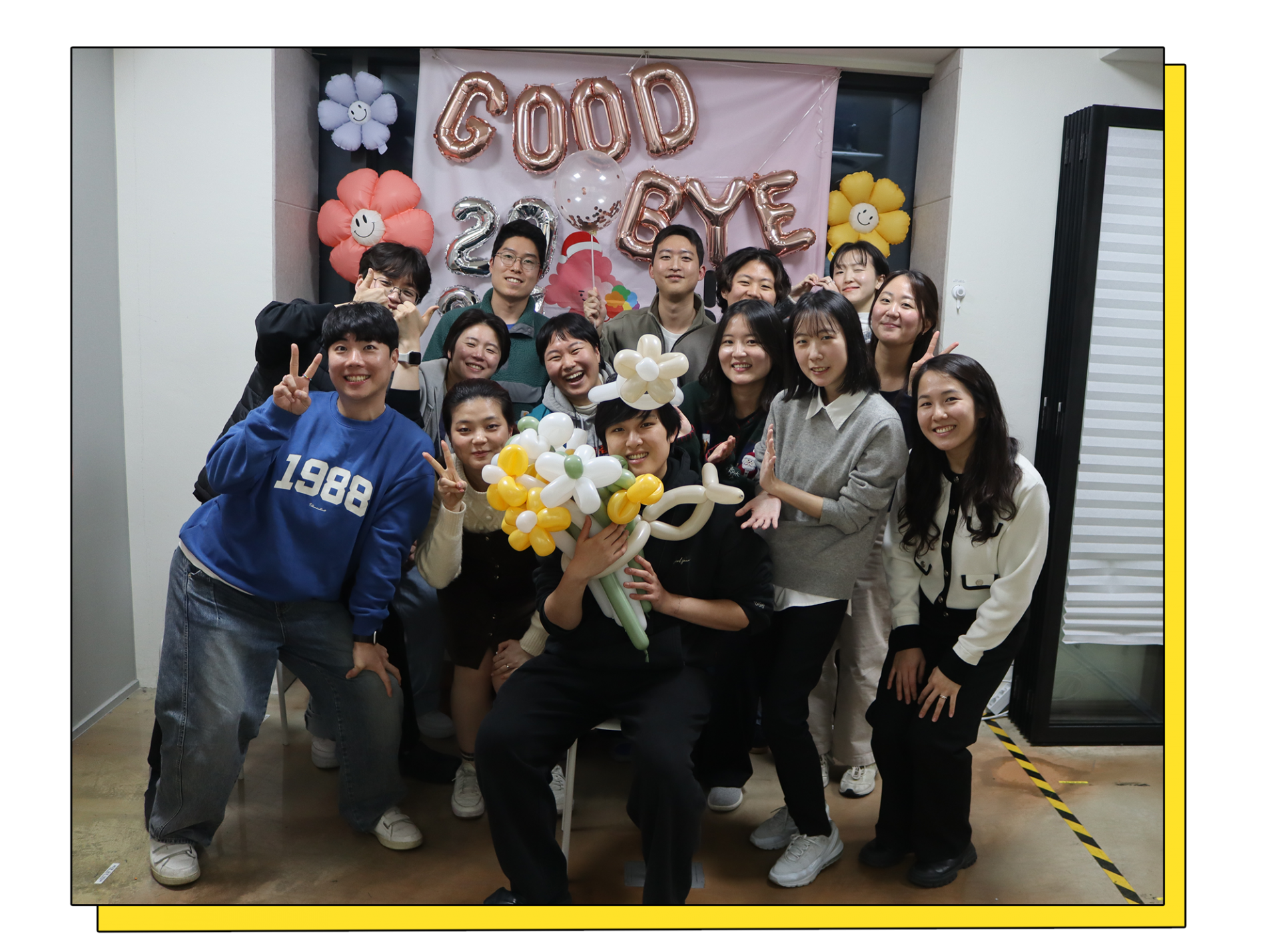 14명의 사람들이 환하게 웃으며 저마다의 포즈를 하고 촬영한 단체사진. 뒤에는 풍선으로 장식이 되어 있고 _GOOD BYE_라고 이니셜 풍선이 붙어 있다