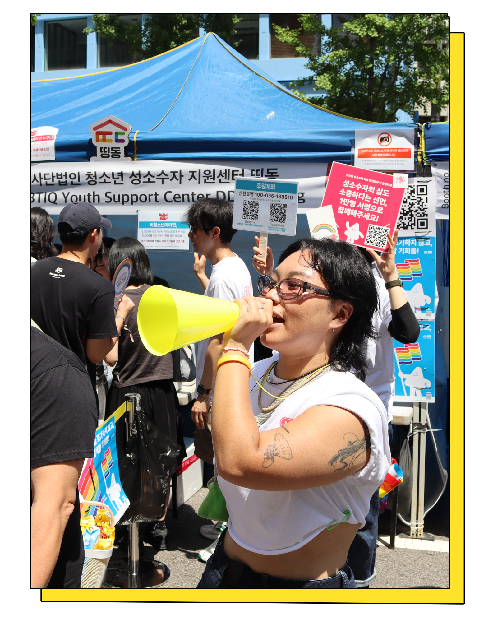 퀴어문화축제의 띵동 부스에 많은 인파가 모여 있고, 그 가운데 한 활동가가 확성기를 한 손에 들고 캠페인을 홍보하고 있다. 활동가 뒤에는 '성소수자의 삶도 소중하다는 선언, 1만
